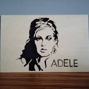 May include: A black and white stencil portrait of Adele on a light-colored wooden plaque. The singer's name, "ADELE," is printed in a simple font below the image. The artwork is displayed on a wooden surface.