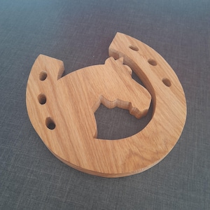 May include: A wooden horseshoe-shaped coaster with a horse head cutout. The coaster is made of light brown wood and features three circular holes on each side of the horseshoe. The horse head is in the center of the horseshoe.