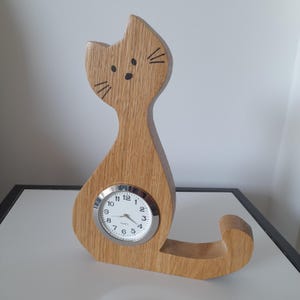 May include: Wooden cat-shaped clock with a round clock face. The clock is made of light brown wood and features black painted whiskers and eyes. The clock face has black numbers and hands.