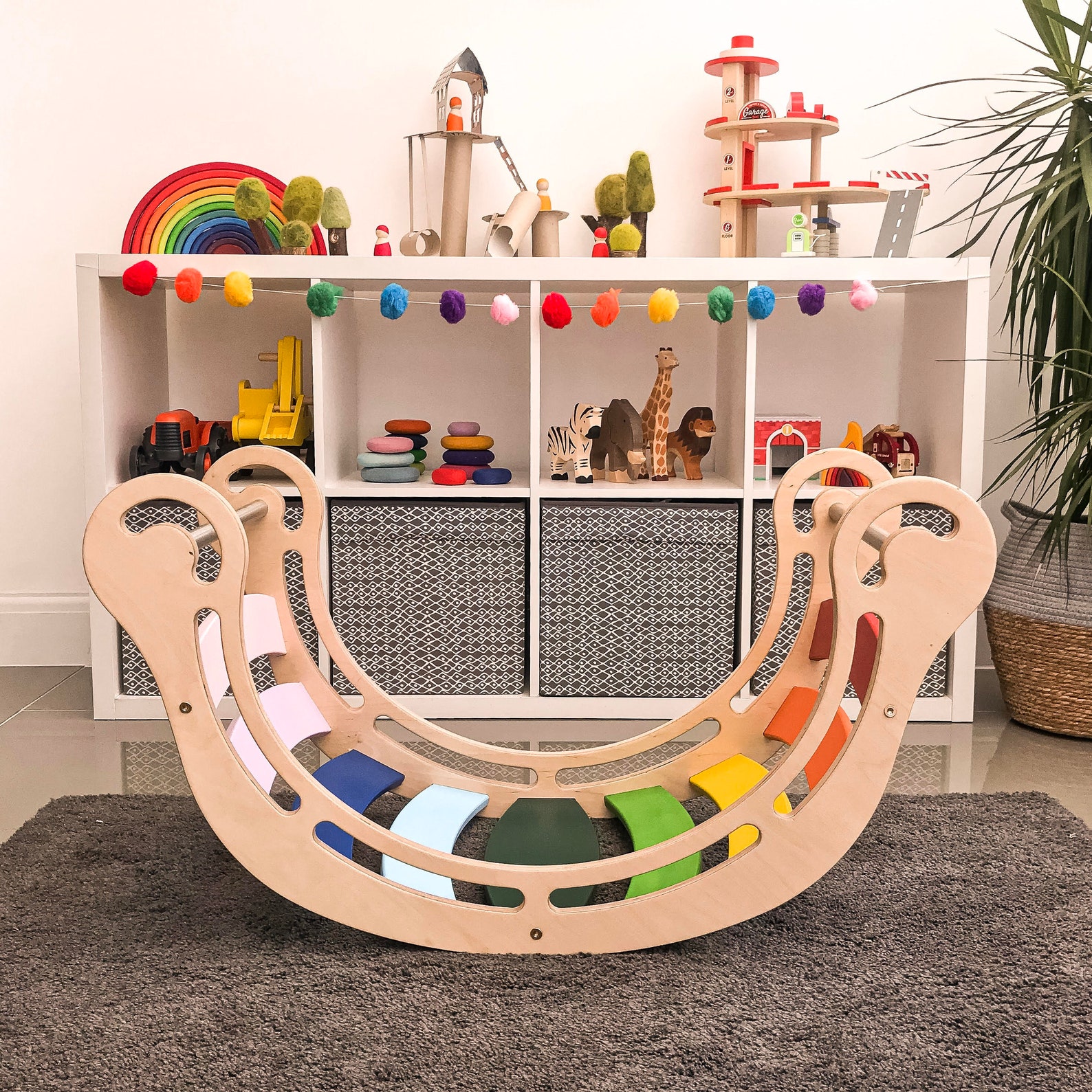 A wooden rocking rainbow on a grey rug in front of toddler shelves featuring wooden toys like a stacking rainbow, wood animals, and staging stones. 