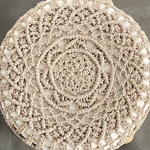 Op de afbeelding: Een rond, beige gebreid kussen met een cirkelvormig patroon. Het kussen is gemaakt van dik, geknoopt garen en heeft een wit centrum.