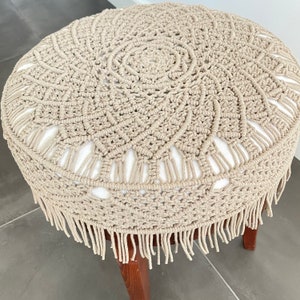 May include: A beige crocheted stool cover with a fringe border. The cover features a circular design with a central circle and a repeating pattern of smaller circles.