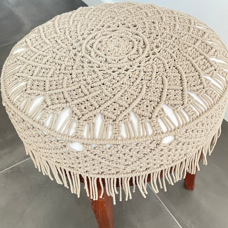May include: A beige crocheted stool cover with a fringe border. The cover features a circular design with a central circle and a repeating pattern of smaller circles.