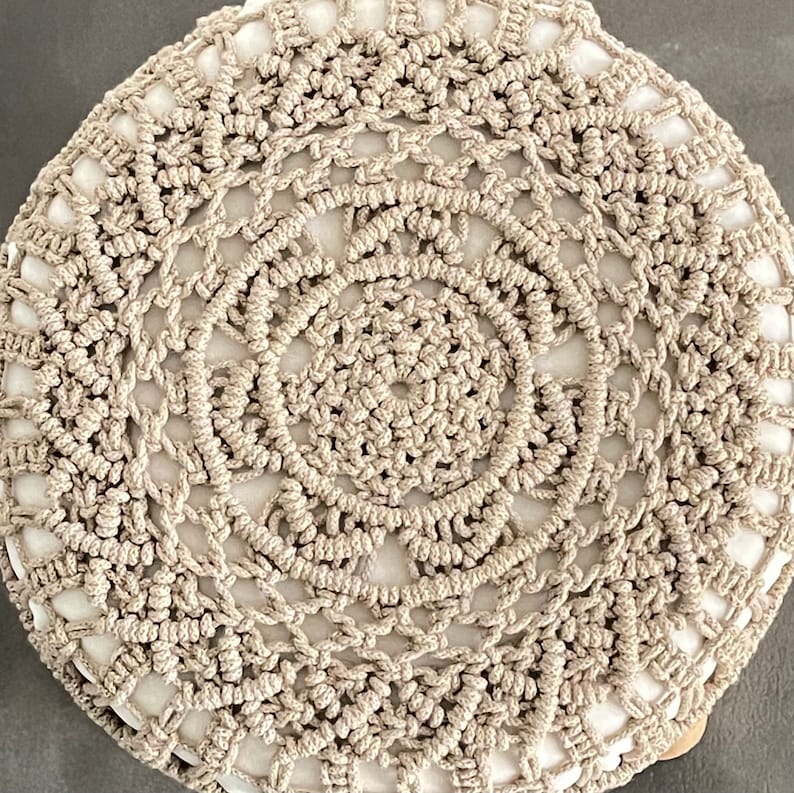 Op de afbeelding: Een rond, beige gebreid kussen met een cirkelvormig patroon. Het kussen is gemaakt van dik, geknoopt garen en heeft een wit centrum.