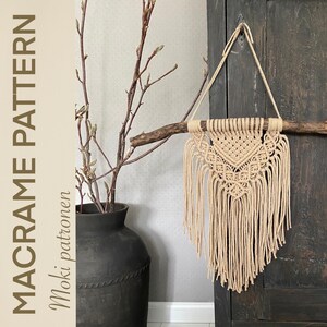 Op de afbeelding: Een beige macramé wandhanger met een geometrisch patroon, hangend aan een tak aan een donkerhouten muur. De macramé is gemaakt van natuurlijke vezelkoord en heeft een franje aan de onderkant.