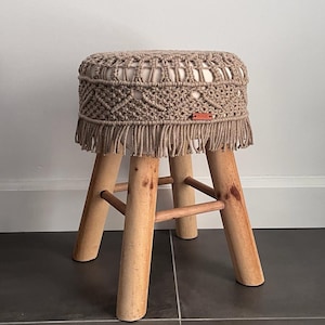 Op de afbeelding: Een beige macram&eacute; kruk met een franje rand en houten poten. De kruk heeft een label met de tekst "boho".