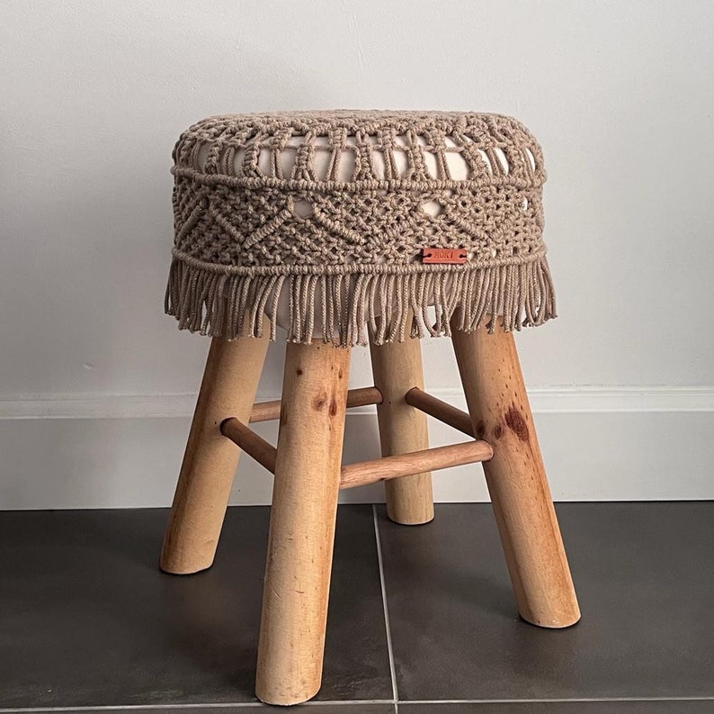 Op de afbeelding: Een beige macram&eacute; kruk met een franje rand en houten poten. De kruk heeft een label met de tekst "boho".