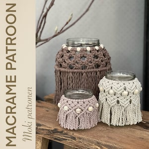 May include: Three macrame candle holders in different sizes, with brown, beige, and light pink cord, and white wooden beads. The candle holders are on a wooden surface.