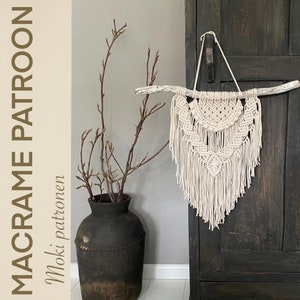 Op de afbeelding: Een witte macramé wandhanger met een geometrisch patroon hangt aan een tak aan een donkerhouten kast. De macramé is gemaakt van dik katoenen koord en heeft een franje aan de onderkant. De kast is donkerbruin van kleur en heeft een verweerde afwerking.