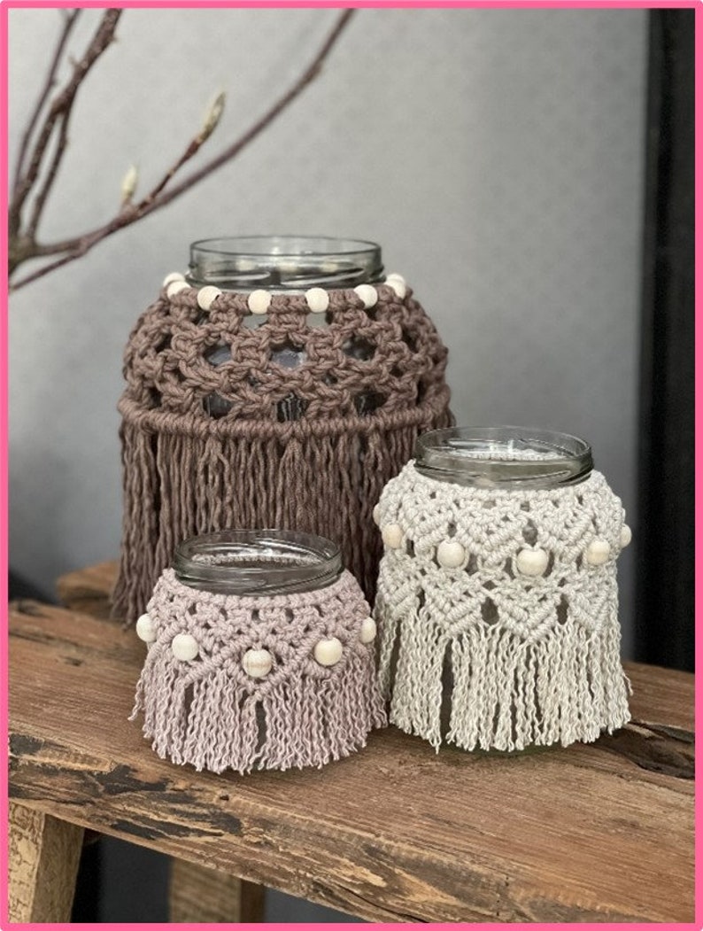 May include: Three glass jars with crocheted covers and white beads. The largest jar has a brown cover with fringe, the medium jar has a white cover with fringe, and the smallest jar has a light pink cover with fringe.