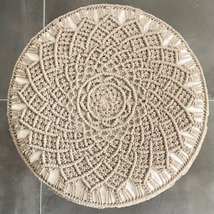 May include: A round, beige macrame rug with a circular pattern in the centre. The rug has a white border.