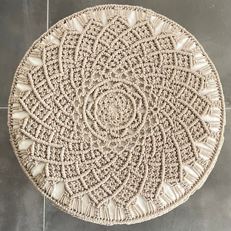 May include: A round, beige macrame rug with a circular pattern in the centre. The rug has a white border.