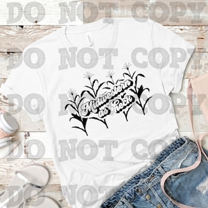 May include: White t-shirt with a black graphic of corn stalks and the text "Midwestern as Heck". The shirt is displayed with denim shorts, a pink purse, and white headphones.