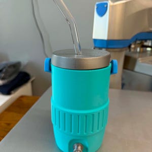May include: A teal insulated tumbler with a silver lid and a clear glass straw. The tumbler has blue handles and a silver spigot at the bottom. The design includes a ribbed section around the middle.