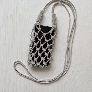 May include: A silver-toned metallic cord phone pouch featuring a knotted net design. The pouch has a long, matching silver cord strap for carrying and contains a solid black rectangular smartphone inside the open-weave structure.