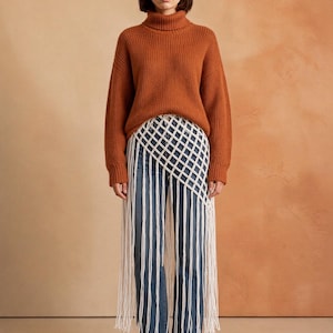 Puede incluir: Un suéter de cuello alto acanalado color óxido combinado con jeans azules y una falda superpuesta de macramé blanco con un patrón de cuadrícula y largos flecos colgantes que llegan hasta los tobillos, usado con botines de gamuza marrón.