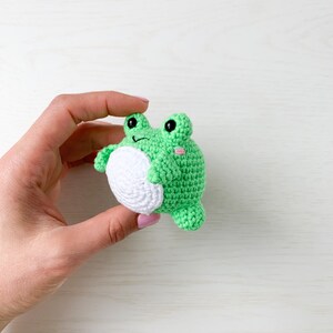 May include: A green crocheted frog toy with white accents and black eyes. The frog is being held in a hand.