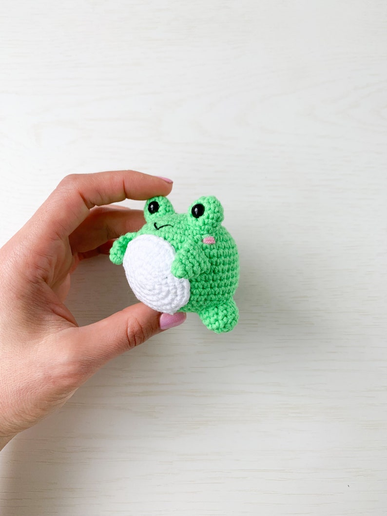 May include: A green crocheted frog toy with white accents and black eyes. The frog is being held in a hand.