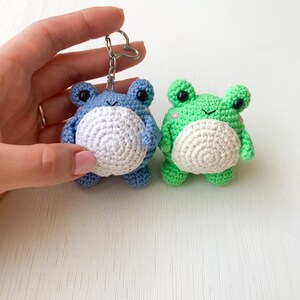 May include: Two crocheted frog keychains, one blue and one green, with white bellies and black eyes. The keychains are hanging from a silver keyring.