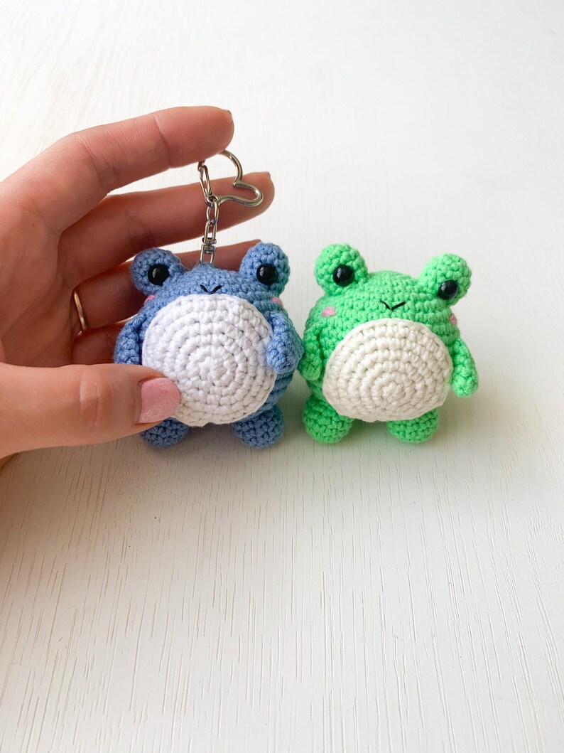 May include: Two crocheted frog keychains, one blue and one green, with white bellies and black eyes. The keychains are hanging from a silver keyring.