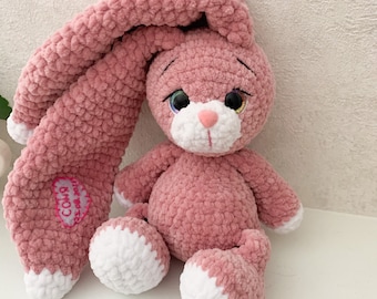 Könnte beinhalten: Ein rosa gehäkeltes Häschen mit weißen Akzenten an seinen Füßen und seinem Gesicht. Das Häschen hat ein langes, schlaffes Ohr, das über seinen Kopf hängt. Das Häschen hat ein rosa Herz mit dem Wort "CO18" darauf gestickt.