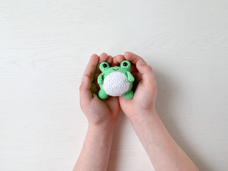 May include: A small green crocheted frog toy with white belly and black eyes, held in the hands of a person.