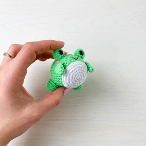 May include: A small green crocheted frog with white accents and black eyes. The frog is being held in a hand.