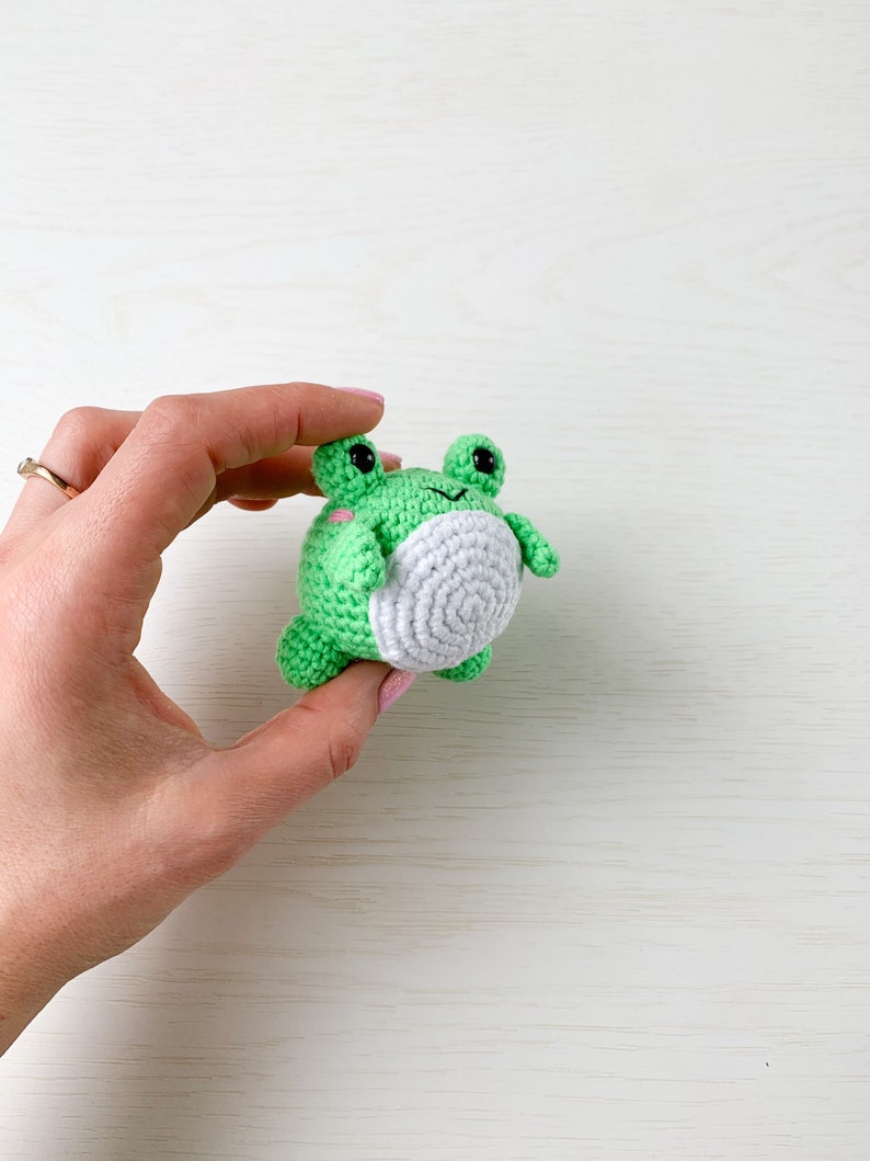 May include: A small green crocheted frog with white accents and black eyes. The frog is being held in a hand.