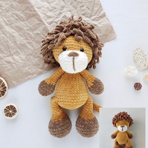 Puede incluir: Un juguete de peluche de león tejido a crochet con pelaje marrón, una melena marrón y rasgos faciales blancos. El juguete está sentado sobre una superficie blanca.