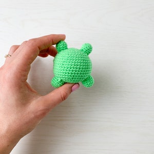 May include: A green crocheted stuffed animal with four small bumps on the back. The animal is being held in a hand.