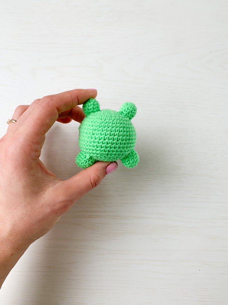 May include: A green crocheted stuffed animal with four small bumps on the back. The animal is being held in a hand.