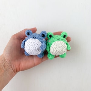 May include: Two crocheted frog plushies, one blue and one green, with white bellies and black eyes. The plushies are held in a hand against a white background.