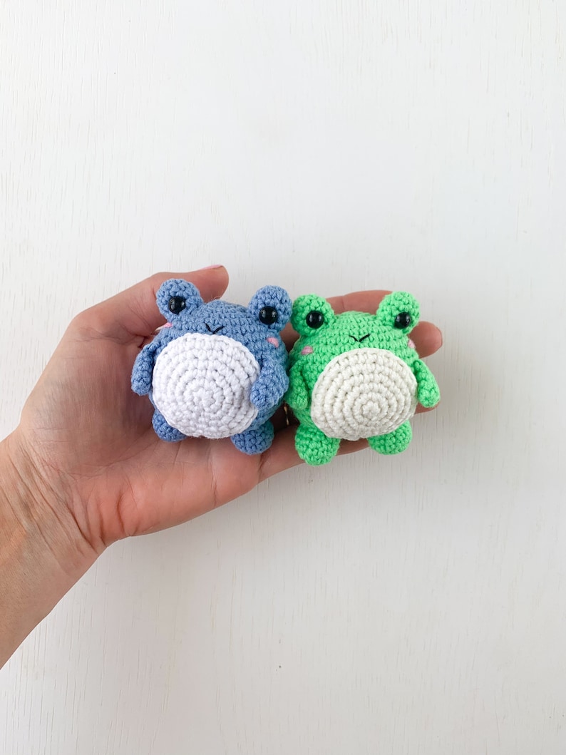 May include: Two crocheted frog plushies, one blue and one green, with white bellies and black eyes. The plushies are held in a hand against a white background.