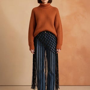 May include: A rust-colored ribbed turtleneck sweater paired with blue denim jeans featuring a black macrame overlay that extends into long, dangling fringe strands, and brown suede ankle boots with a block heel.