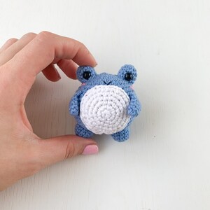 May include: A small crocheted blue frog with white belly and black eyes. The frog is being held by a hand.