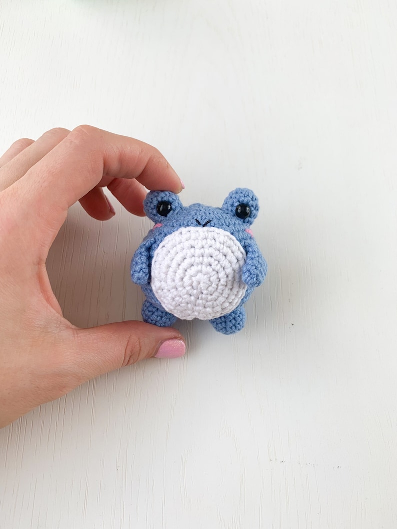 May include: A small crocheted blue frog with white belly and black eyes. The frog is being held by a hand.