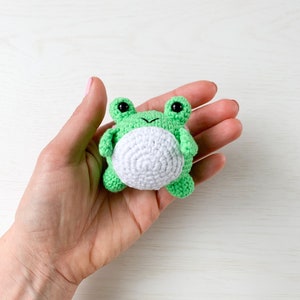May include: A small green crocheted frog with white belly and black eyes. The frog is being held in a hand.