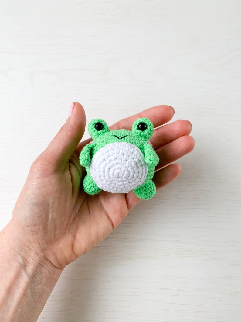 May include: A small green crocheted frog with white belly and black eyes. The frog is being held in a hand.