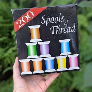May include: A black square box labeled 200 Spools of Thread in white text with a red diagonal stripe. The box features a graphic illustration of a pyramid of colorful thread spools in shades of orange, green, purple, blue, yellow, and pink.
