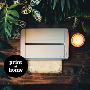Op de afbeelding: Een witte printer met een vel papier op de bovenkant. De printer staat op een houten oppervlak met een brandende kaars en groene planten op de achtergrond. De tekst "print at home" staat op een zwarte cirkel.
