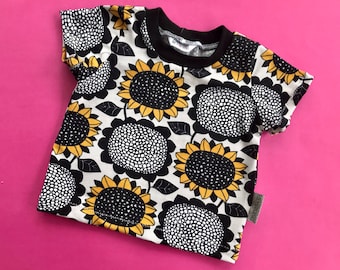 May include: A short-sleeved t-shirt with a repeating sunflower pattern. The design features large black and white sunflowers with yellow centres and green leaves, set against an off-white background. The shirt has a black collar.