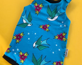 Puede incluir: Un body de bebé azul con un estampado de golondrinas y flores. El body tiene un ribete negro y cierres de botón a presión en los hombros. El diseño incluye estrellas blancas y flores rosas.