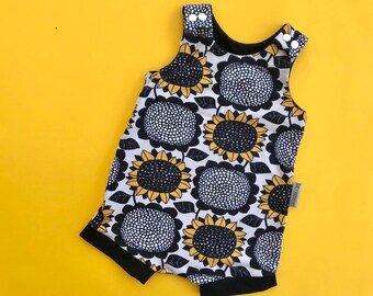 Puede incluir: Un body sin mangas para bebé con un estampado de girasoles. El body presenta un fondo blanco con grandes girasoles negros y amarillos. El ribete es negro y el body tiene cierres de botón a presión blancos en los hombros.