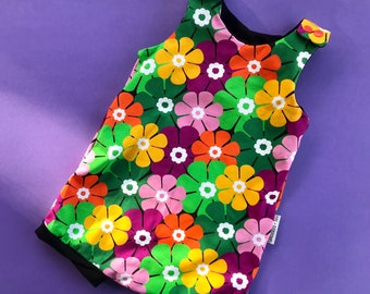 Puede incluir: Prenda sin mangas con un vibrante estampado floral en tonos verdes, amarillos, naranjas, rosas y morados. La prenda tiene un ribete negro y está sobre un fondo morado. El diseño floral presenta detalles blancos.