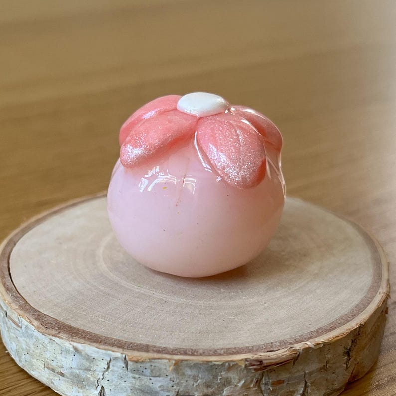 Pu&ograve; includere: Un oggetto decorativo a forma di fiore in vetro rosa con un centro bianco. I petali hanno una finitura glitterata. L'oggetto &egrave; appoggiato su una piccola fetta di legno rotonda.