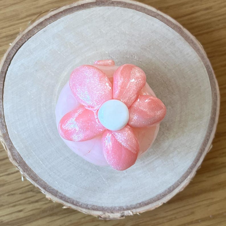 Pu&ograve; includere: Un oggetto a forma di fiore rosa con un centro bianco, possibilmente un oggetto decorativo o un accessorio. Il fiore ha cinque petali con accenti glitterati. L'oggetto &egrave; posto su una fetta di legno chiaro, creando un contrasto naturale.