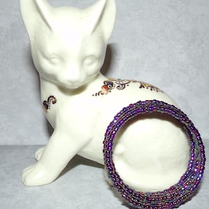 May include: A multi-strand bracelet made of iridescent purple beads. The bracelet is coiled and lying on a white porcelain figurine of a cat.