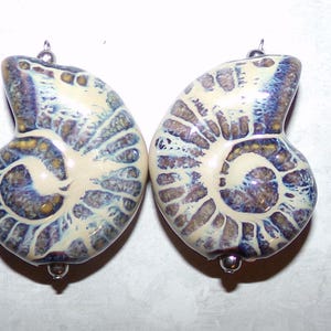 Op de afbeelding: Twee witte keramische ammoniet schelpvormige kralen met bruine en paarse swirls. De kralen hebben zilveren metalen lussen om op te hangen.