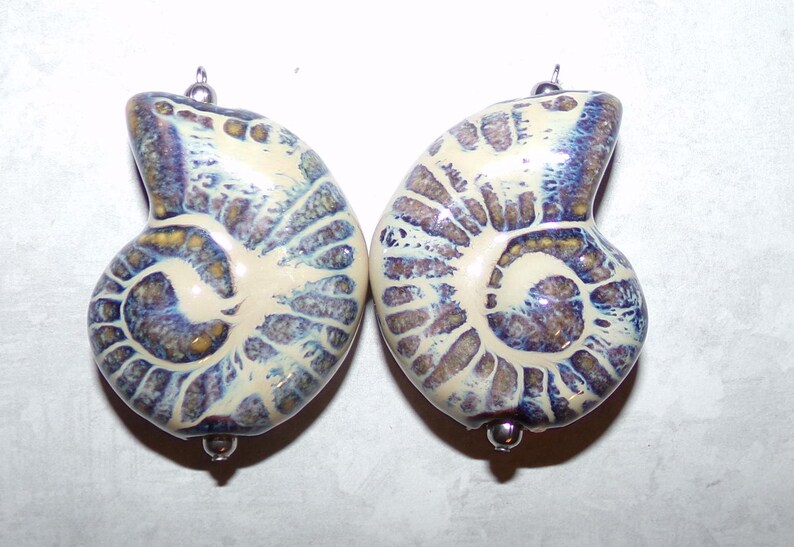 Op de afbeelding: Twee witte keramische ammoniet schelpvormige kralen met bruine en paarse swirls. De kralen hebben zilveren metalen lussen om op te hangen.