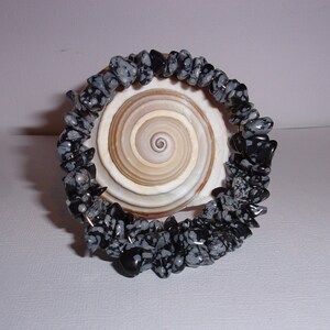 May include: A black and white snowflake obsidian chip bracelet. The bracelet is made of small, irregularly shaped stones strung together. The bracelet is resting on a large, light brown seashell with a spiral pattern.